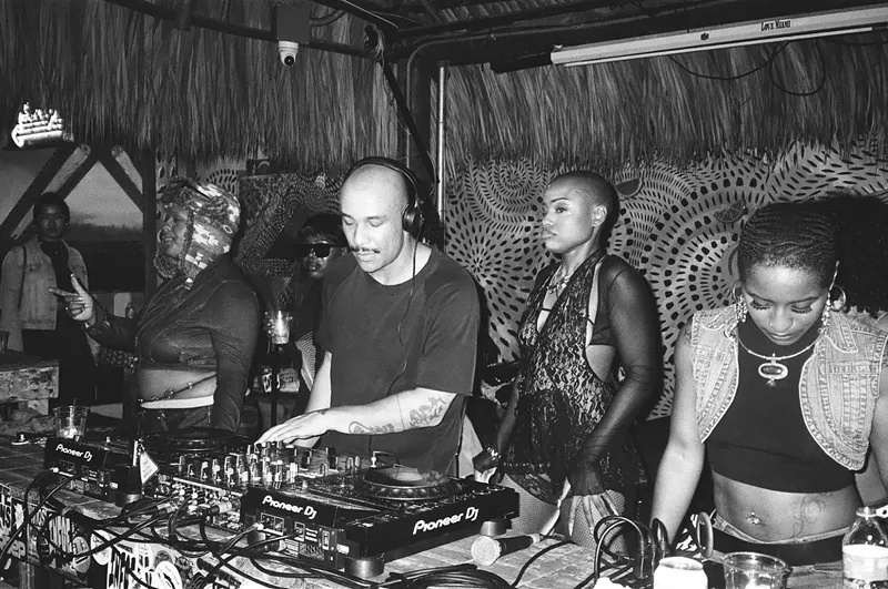 Image: black and white photo of a DJ at the turntables, flanked by several women