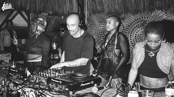 black and white photo of a DJ at the turntables, flanked by several women
