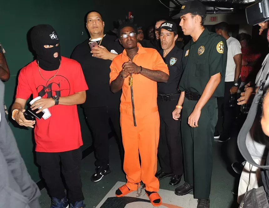 Kodak Black in a posed color shot, dressed in an orange prison jumpsuit, standing with others, including two white men in police gear