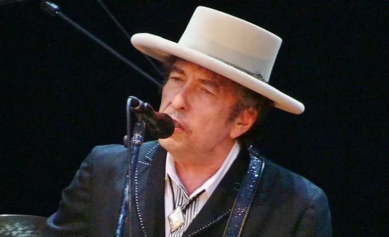Image: headshot of an older bob dylan performing, sporting a wide-brimmed hat